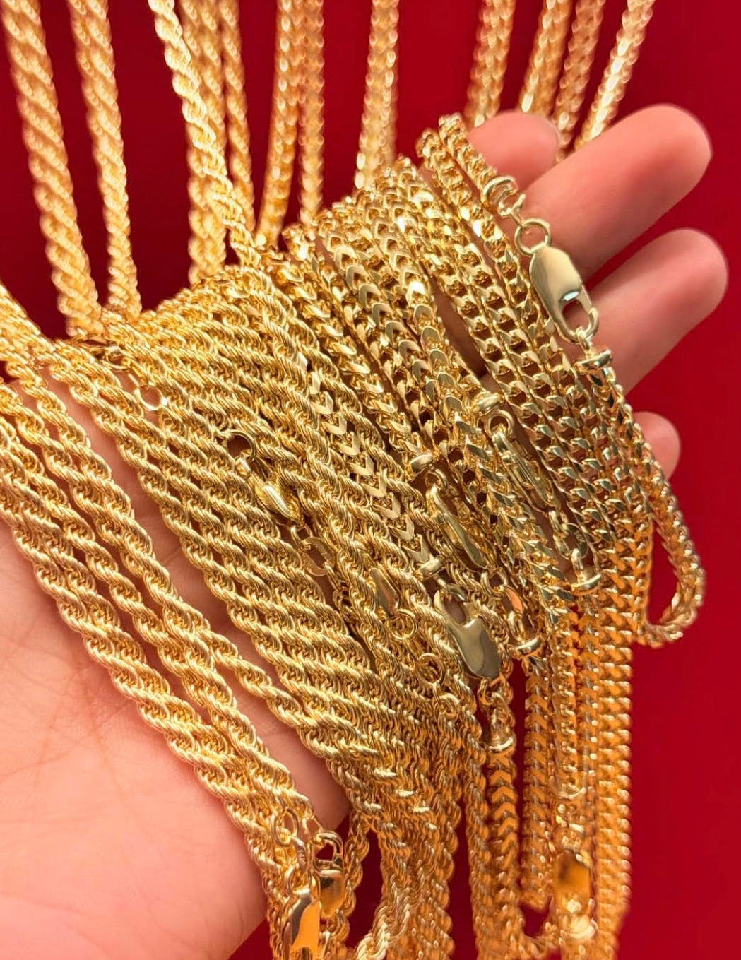A hand holding a bundle of thick gold-toned chain necklaces against a red background.